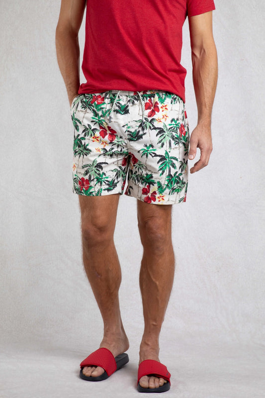 A pair of men's swim shorts with a tropical palm tree and floral pattern, worn with a red top and red slippers.