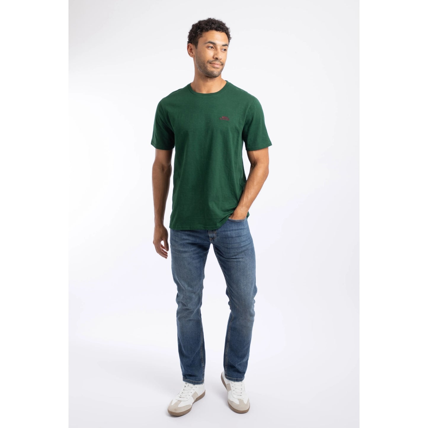 A man wearing a Weird Fish short sleeve Fished t-shirt in Pine Green, with blue jeans and white trainers.
