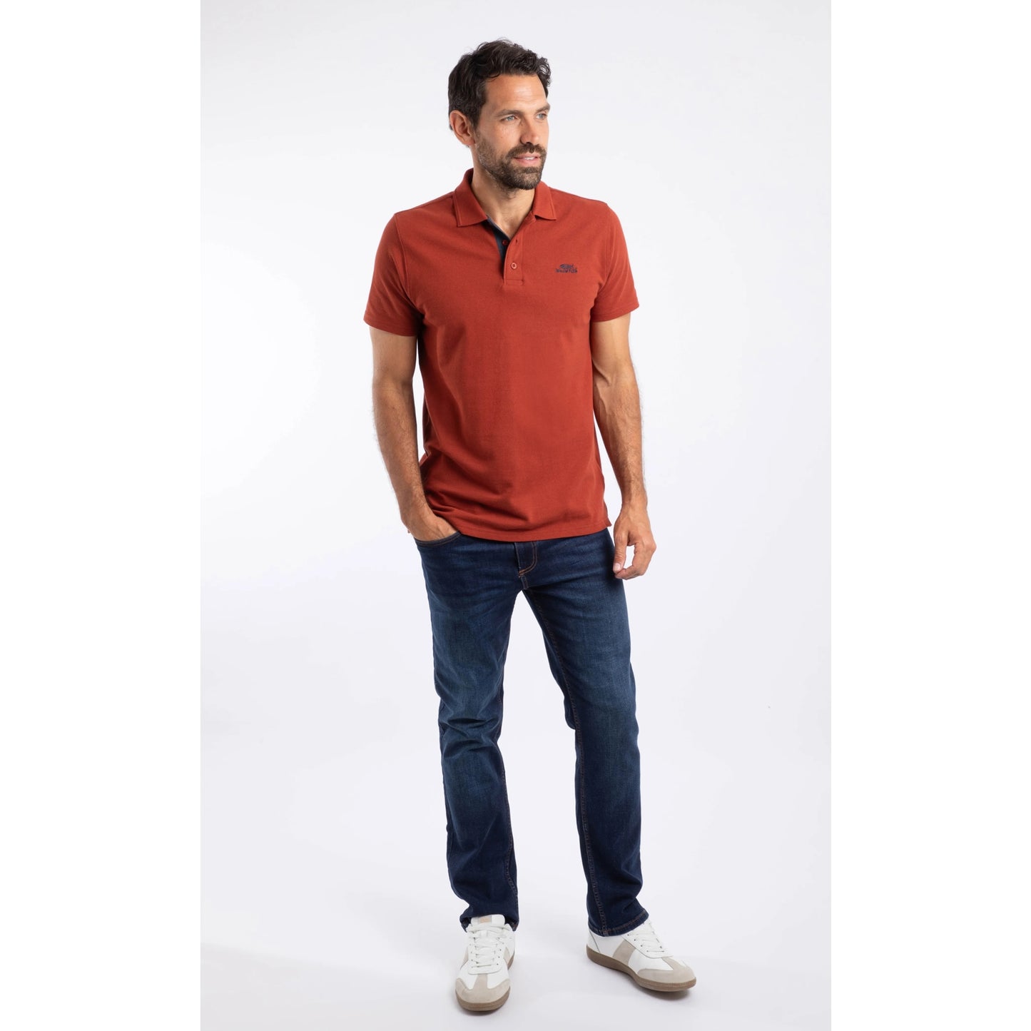 A man wearing a short sleeve Weird Fish Miles polo shirt in Burnt Henna with blue jeans and white trainers.