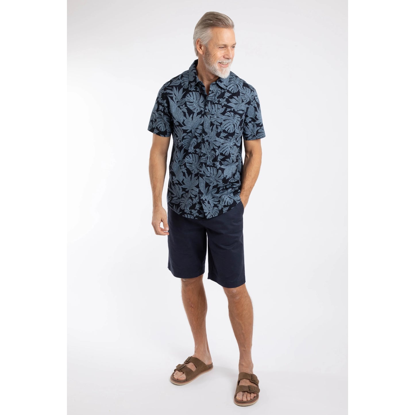 Man wearing a Weird Fish short sleeve Faraway shirt in Blue Mirage with a tropical leaf pattern with navy shorts and brown sandals