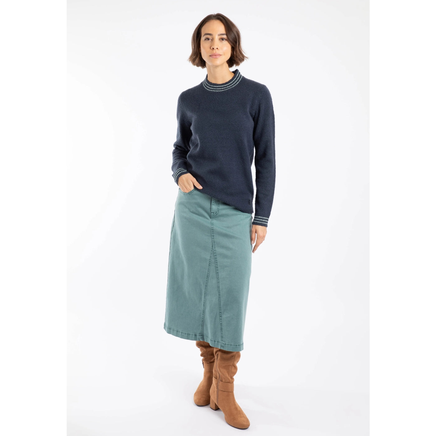 Woman wearing a Weird Fish navy knitted Dawkins jumper, teal skirt, and brown boots on a white background