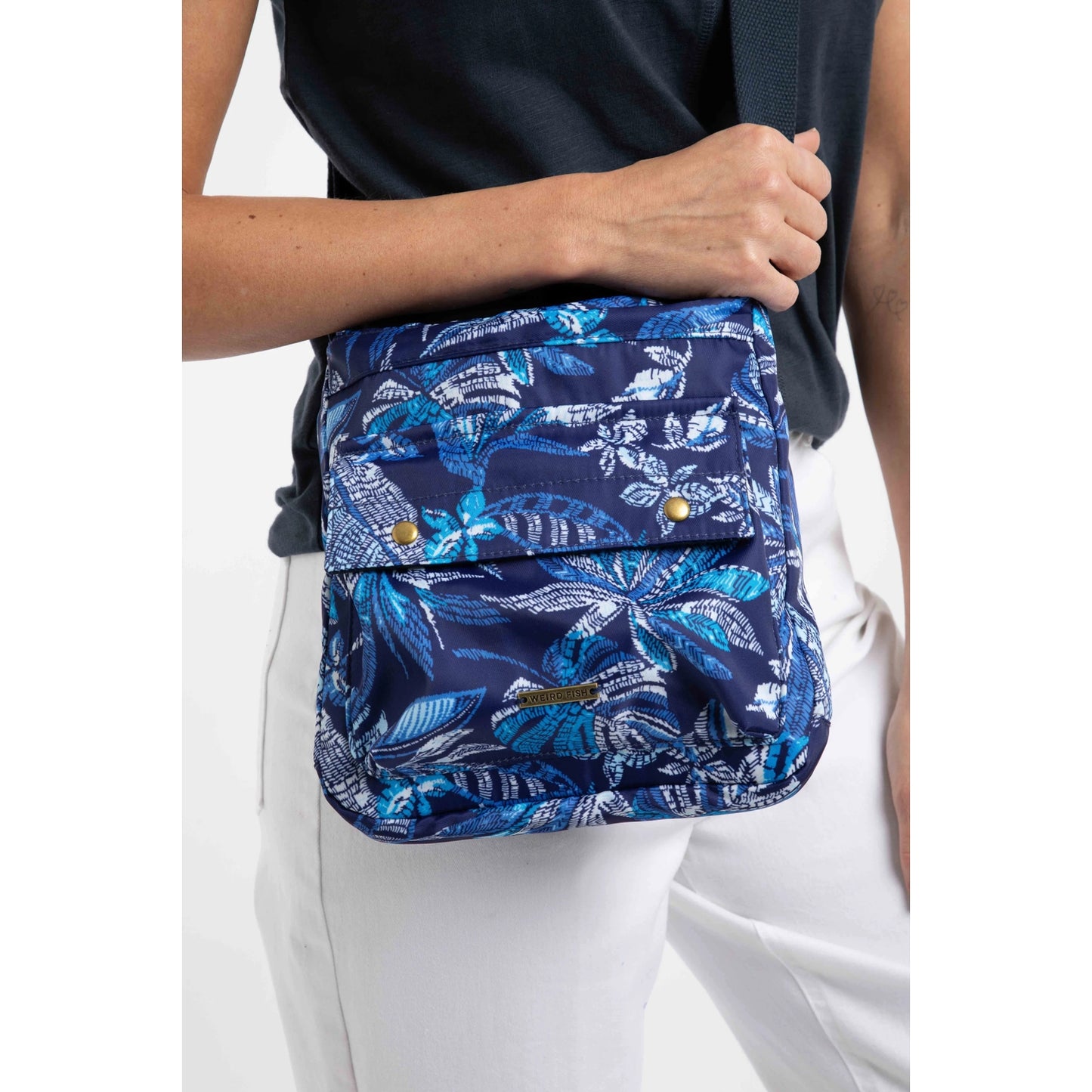 Front view of a woman carrying a Weird Fish Kait cross body bag in navy with a blue tropical leaf print, adjustable shoulder strap and front pocket.