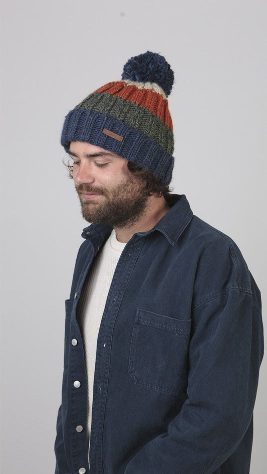 Man wearing a Barts Wilhelm beanie in a stripe multicolour pattern with bobble on top, wearing a navy shirt.
