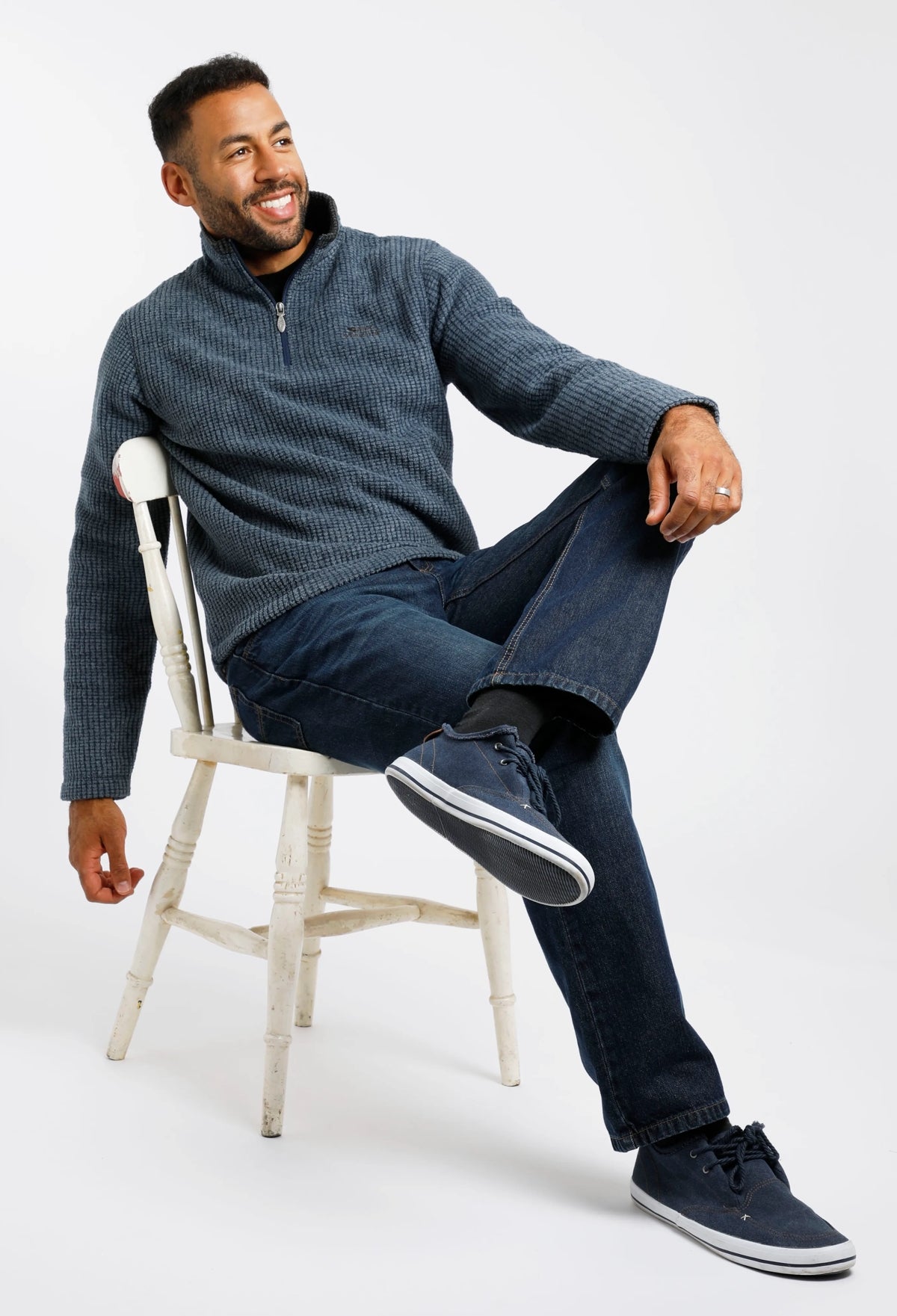 Model wearing Weird Fish Newark fleece in blue, sitting on a white chair with casual jeans and shoes