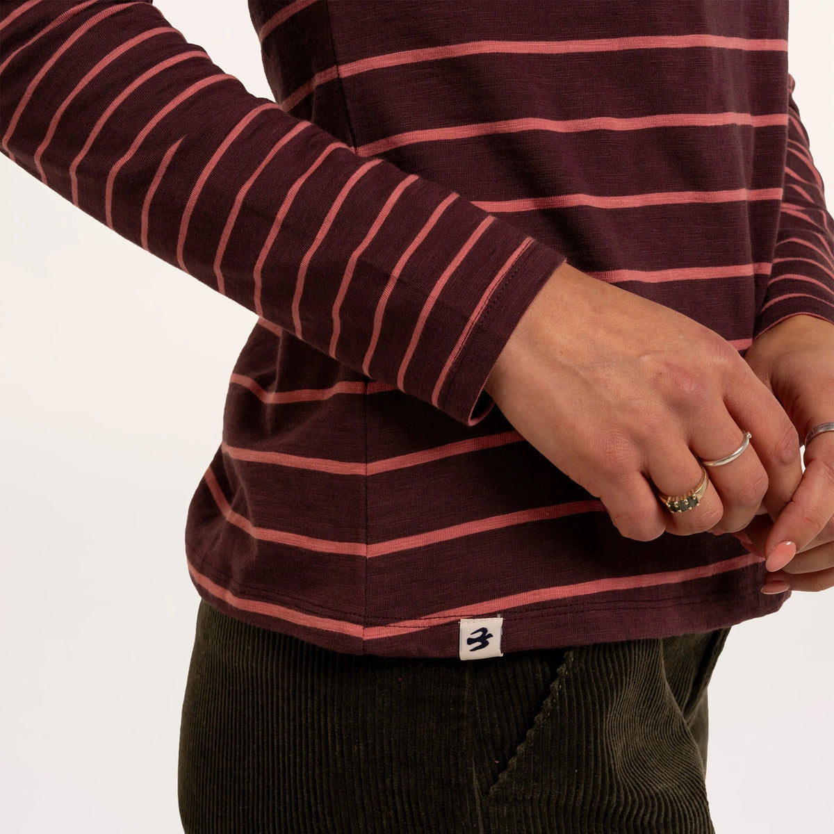 Close-up view of the sleeve and bottom hem of a woman wearing a Brakeburn long sleeve thick and thin stripe Branksome t-shirt in burgundy with coral stripes.
