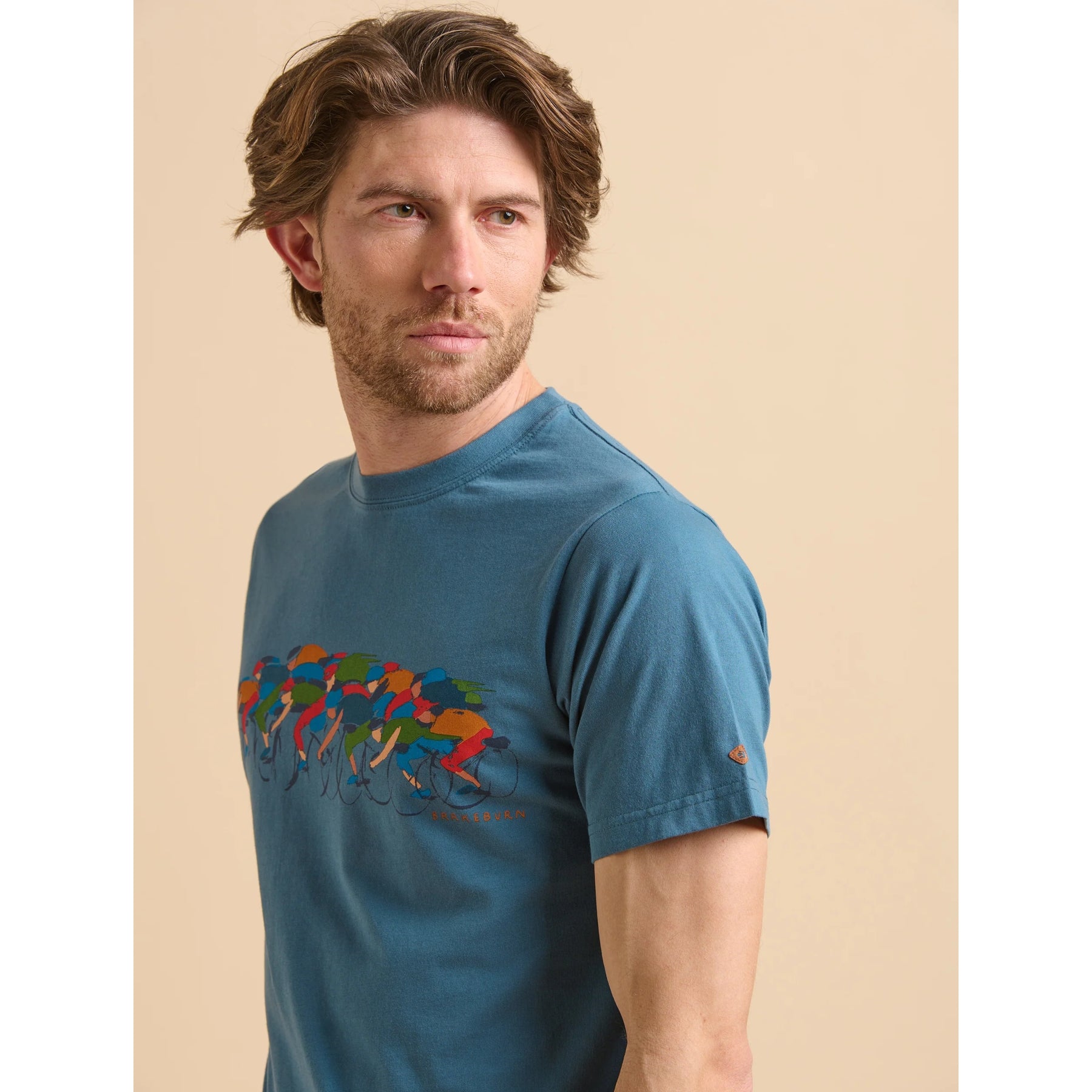 Close-up of a man wearing a Brakeburn blue t-shirt with colourful cycling print on a beige background