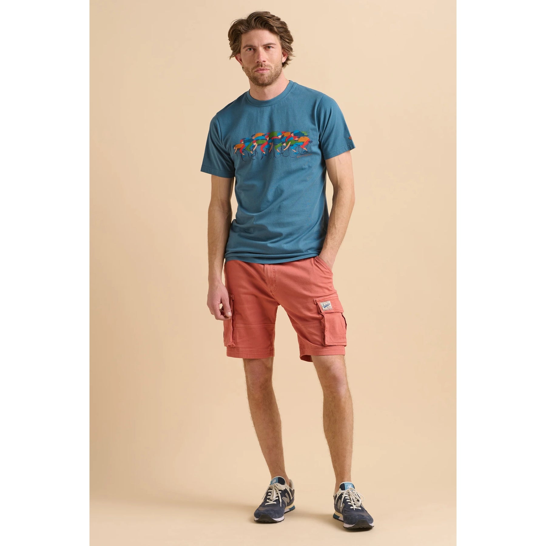 Man wearing a blue Brakeburn t-shirt with a colourful cycling print design and red shorts on a beige background