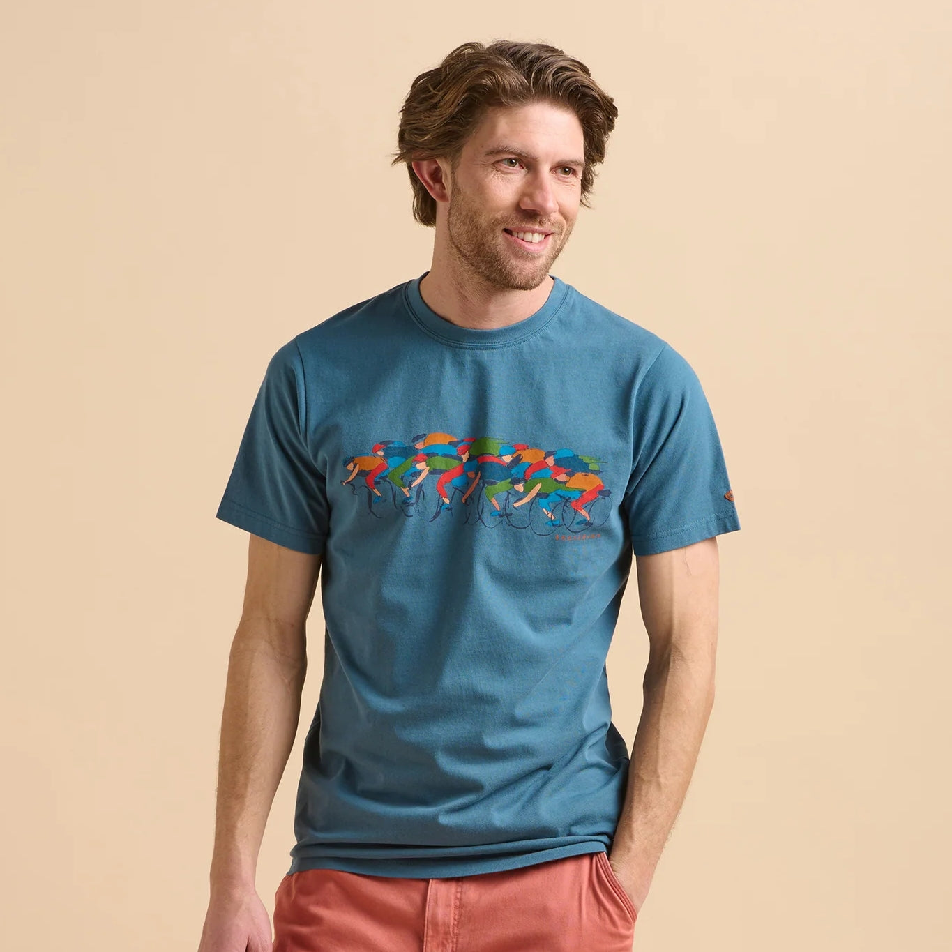 Front view of a man wearing a Brakeburn blue t-shirt with colourful Painted Peleton cyclist print design on a beige background