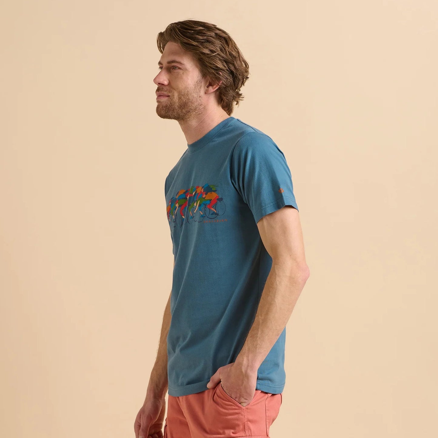 Side view of a man wearing a Brakeburn blue t-shirt with multicolourd Paited Peleton print design on a beige background