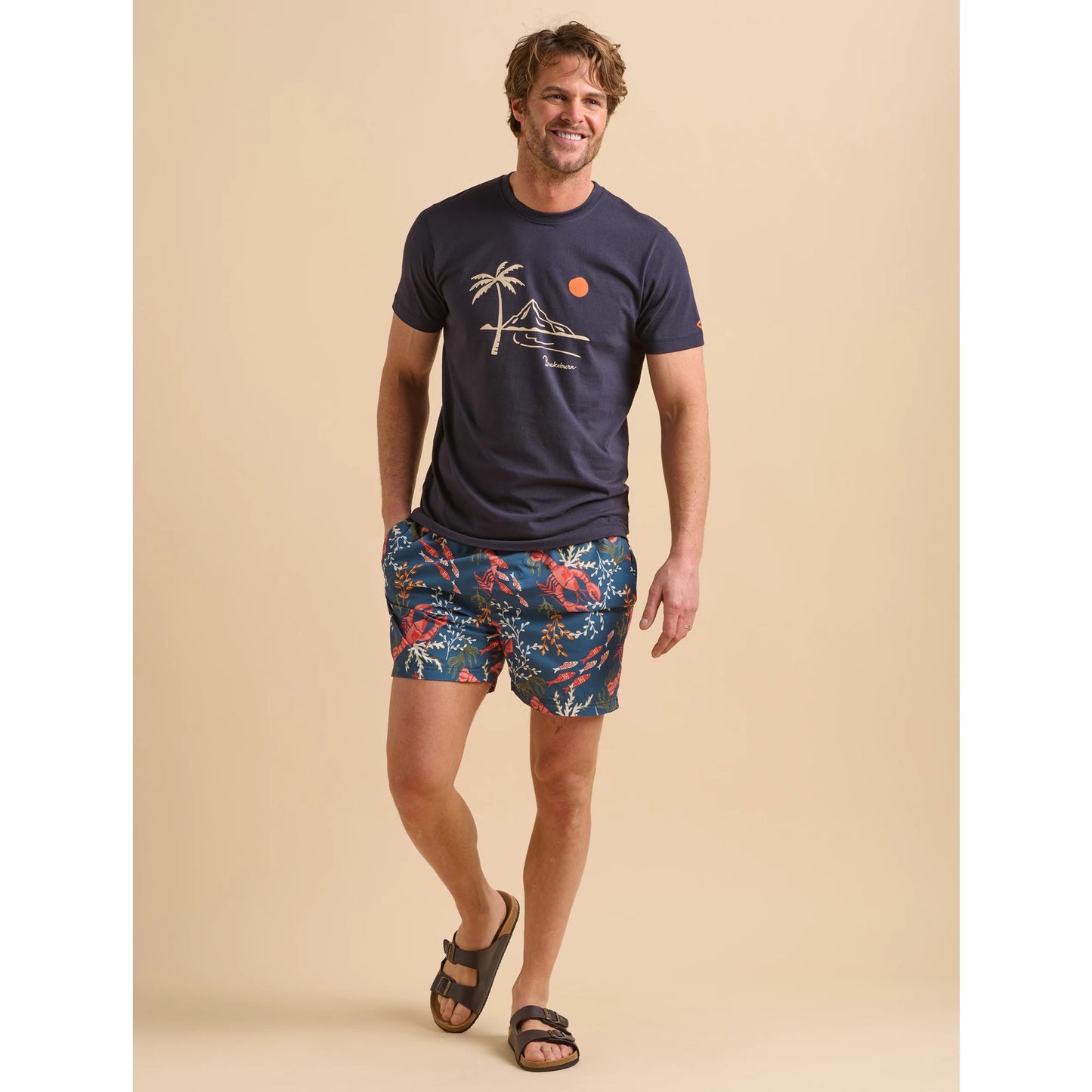 Man wearing a navy Brakeburn Island print t-shirt and colorful shorts on a beige background