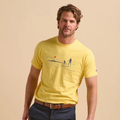 Front view of a man wearing a Brakeburn yellow t-shirt with a South Coast Surf print design on a beige background