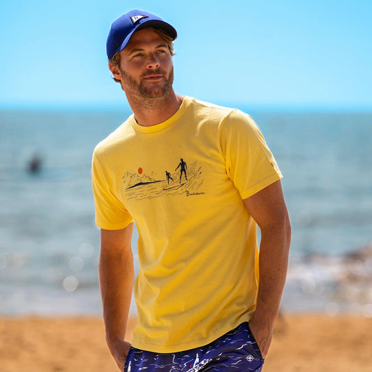 Front view of a man wearing a Brakeburn yellow t-shirt with a South Coast Surf print design on a beach