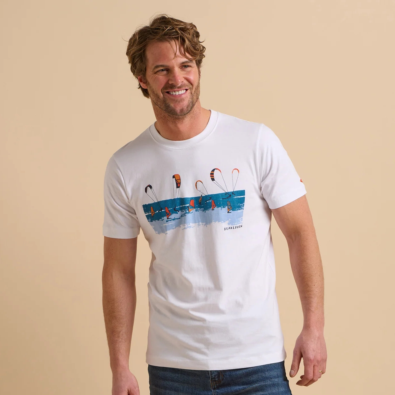 Front view of a man wearing a Brakeburn short sleeve white t-shirt with a windsurfer and kite surfer print design on a beige background