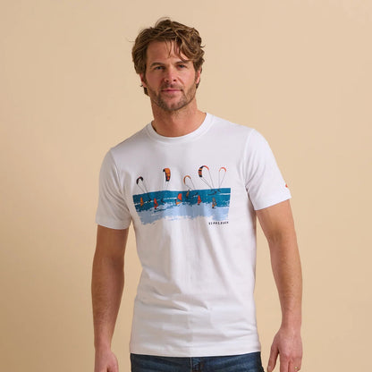 Front view of a man wearing a white short sleeve Brakebun tee with a sea, kite and windsurfer graphic design on a beige background