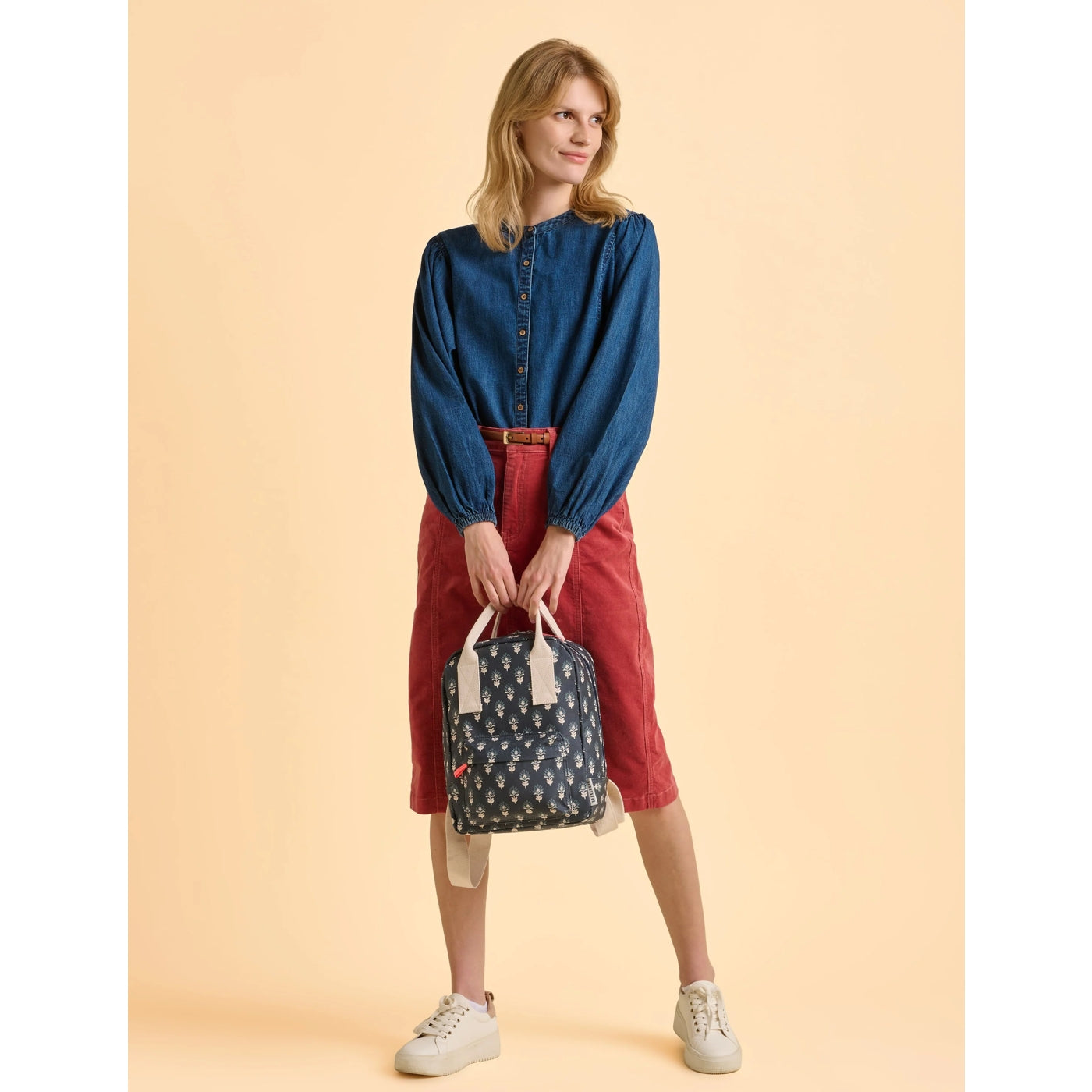 Woman holding a Brakeburn Henna floral patterned navy backpack against a beige background