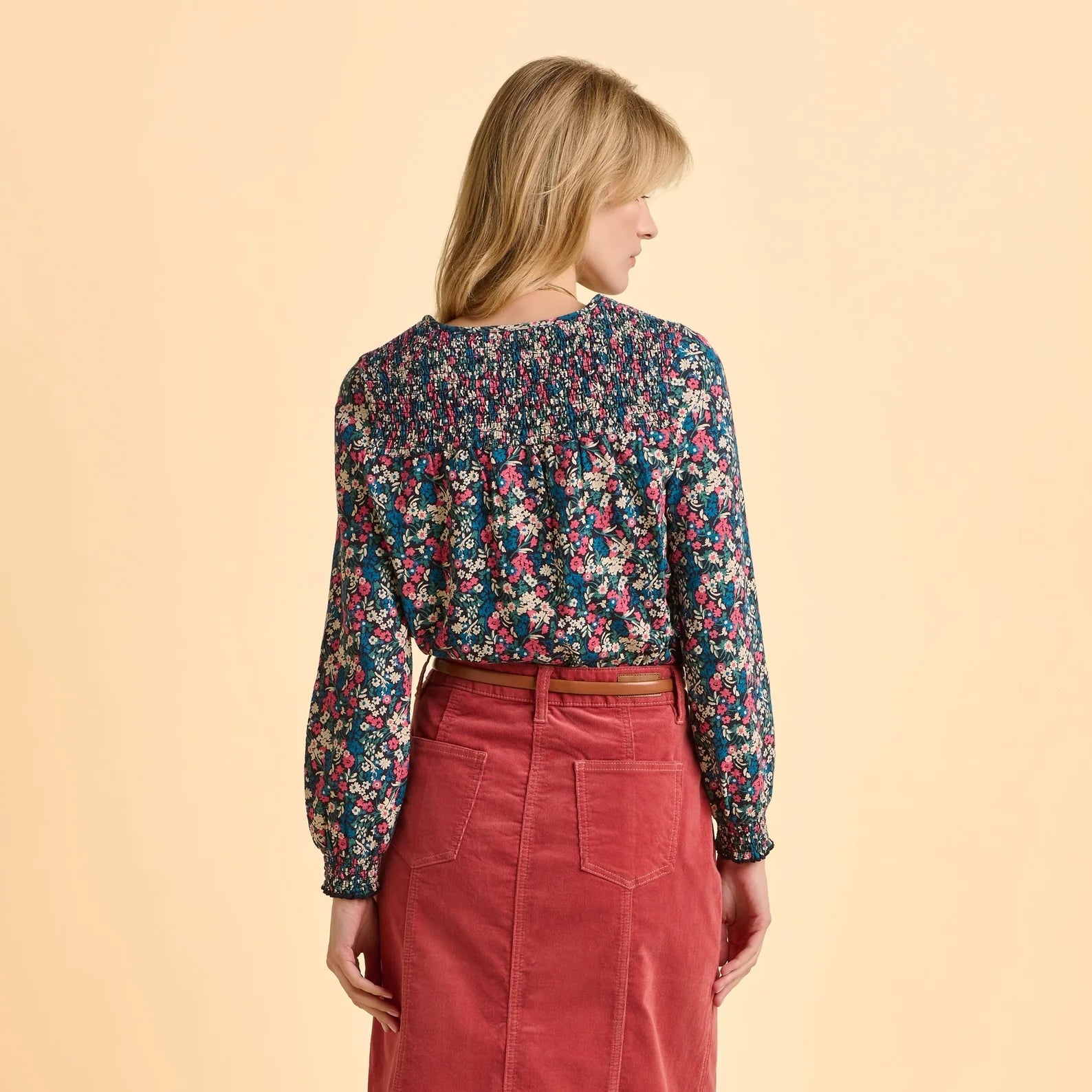 Back view of a woman wearing a Brakeburn Brenda floral long sleeve t-shirt and red skirt on a beige background