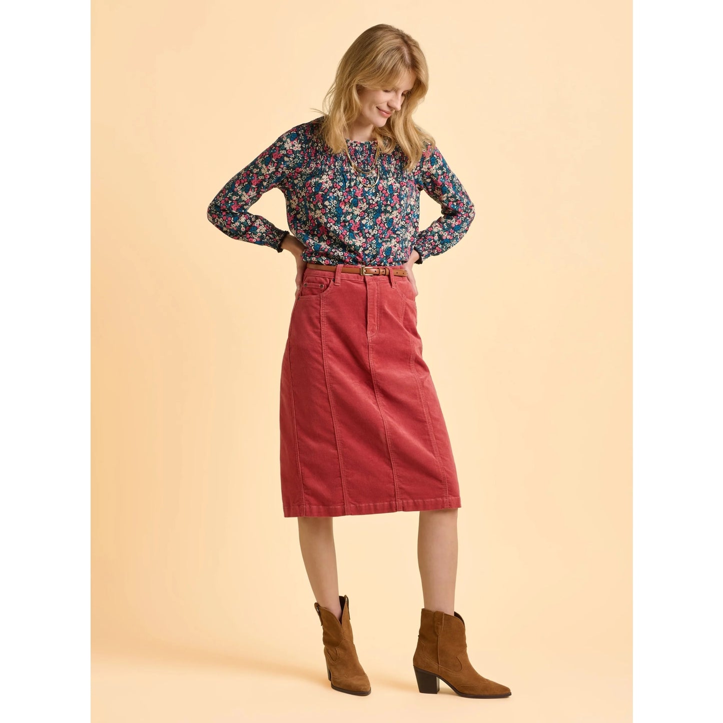 Woman wearing a floral Brakeburn long sleeve floral t-shirt, red skirt, and brown boots on a beige background