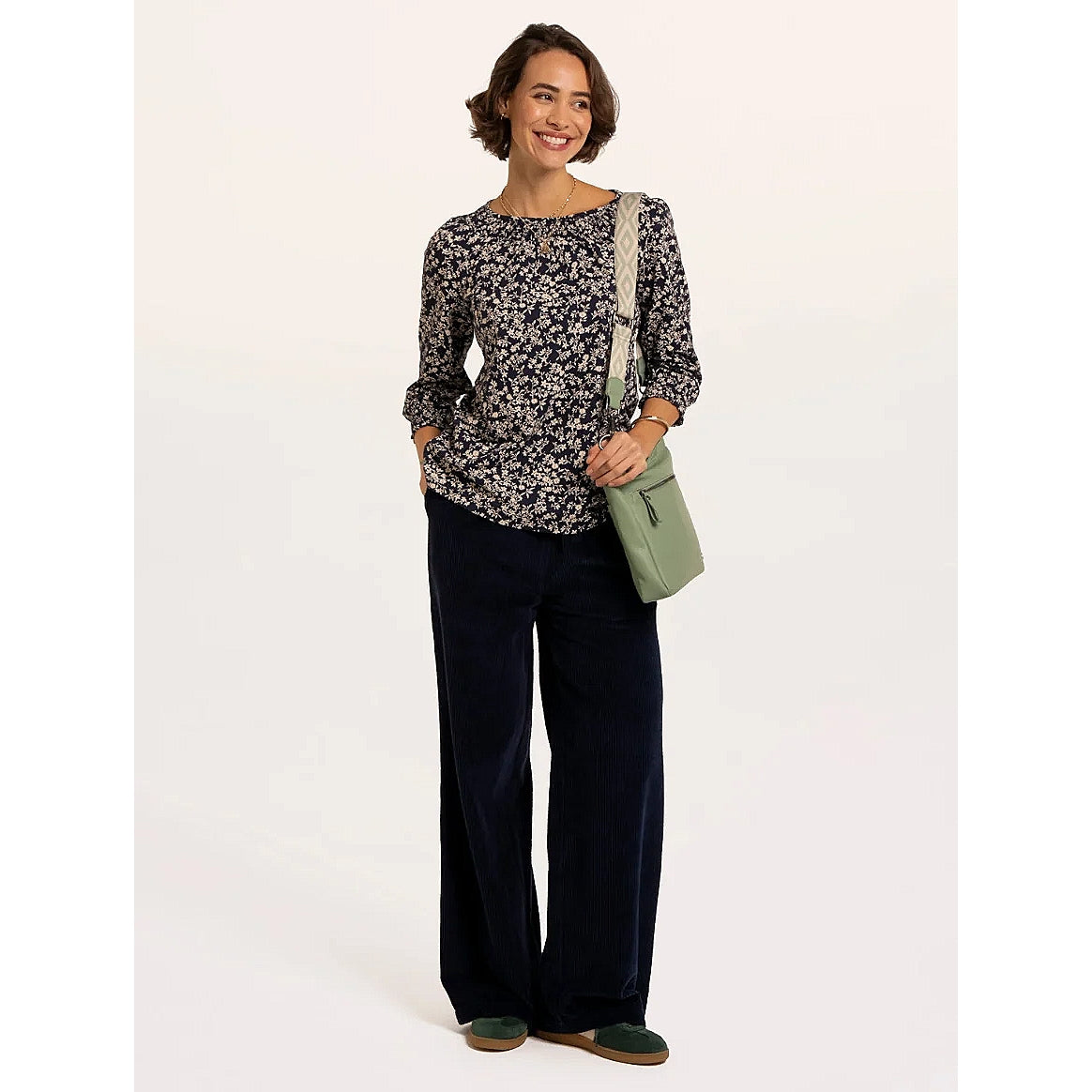 A woman wearing a Brakeburn Brielle 3/4 sleeve t-shirt in navy with a white floral pattern and elasticated cuffs and neckline trim with dark trousers and a green bag.