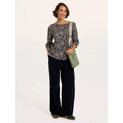 A woman wearing a Brakeburn Brielle 3/4 sleeve t-shirt in navy with a white floral pattern and elasticated cuffs and neckline trim with dark trousers and a green bag.