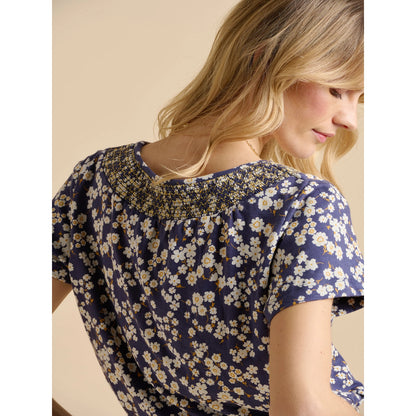 Back view of a woman wearing a Brakeburn short sleeve Floating Daisy t-shirt in navy blue with a white floral pattern, with a neutral background
