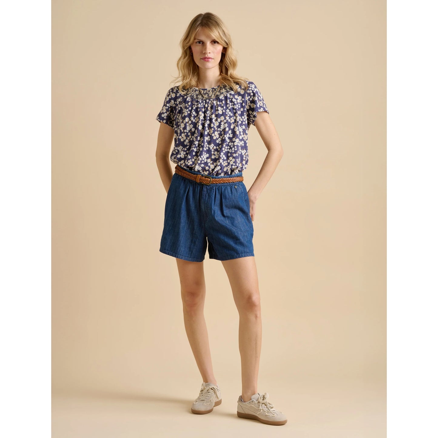 Woman wearing a Brakeburn floral top and blue shorts on a beige background