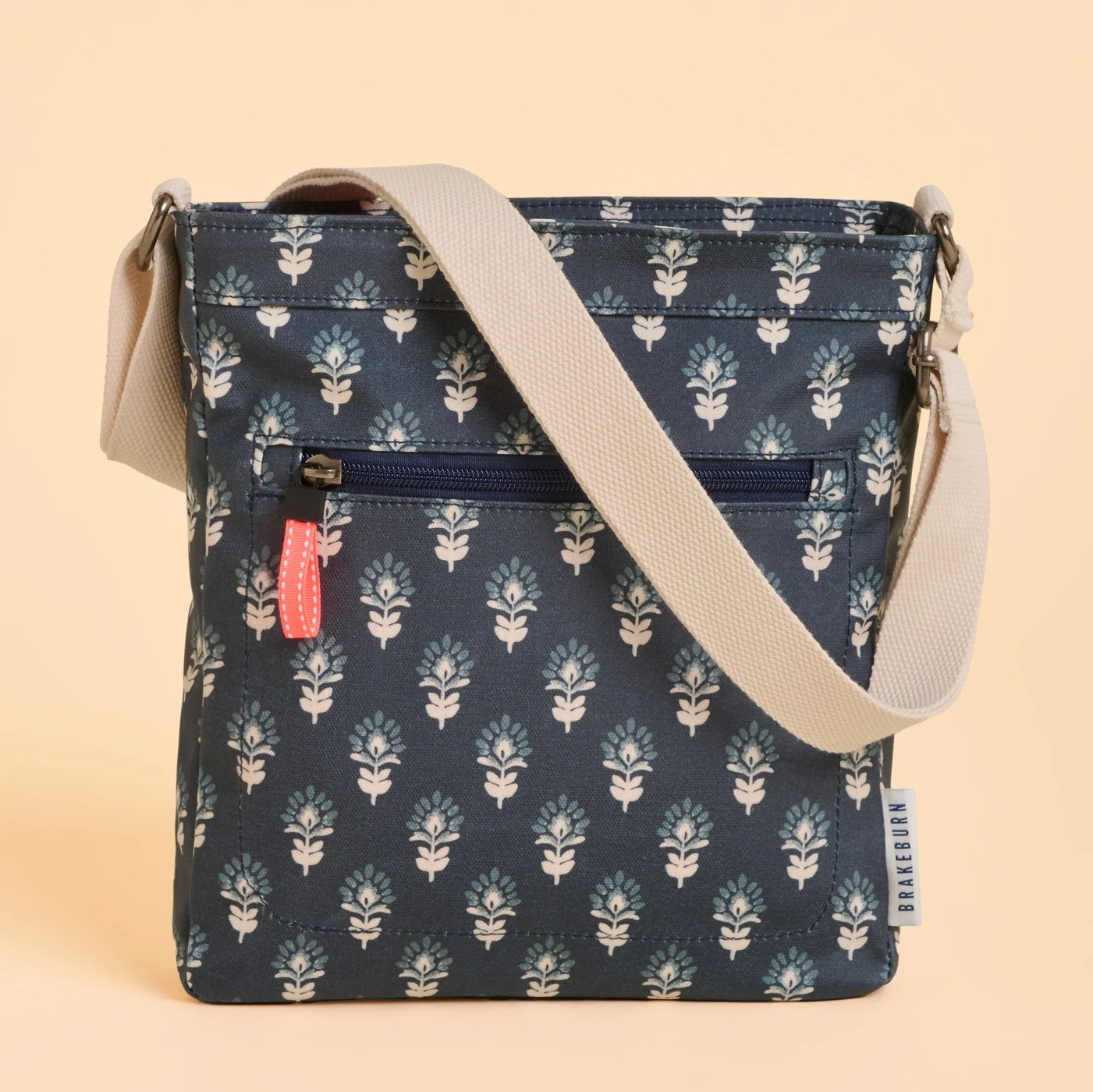 Front view of a Brakeburn Henna floral patterned cross body bag in navy with a beige strap on a beige background