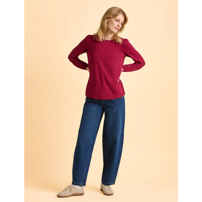 Woman wearing a Brakeburn burgundy red Janis long sleeve t-shirt and blue jeans on a beige background