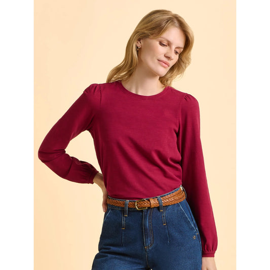 Woman wearing a Brakeburn long sleeve Janis t-shirt in Burgundy Red with gathered shoulder detail, with blue jeans on a cream colour background