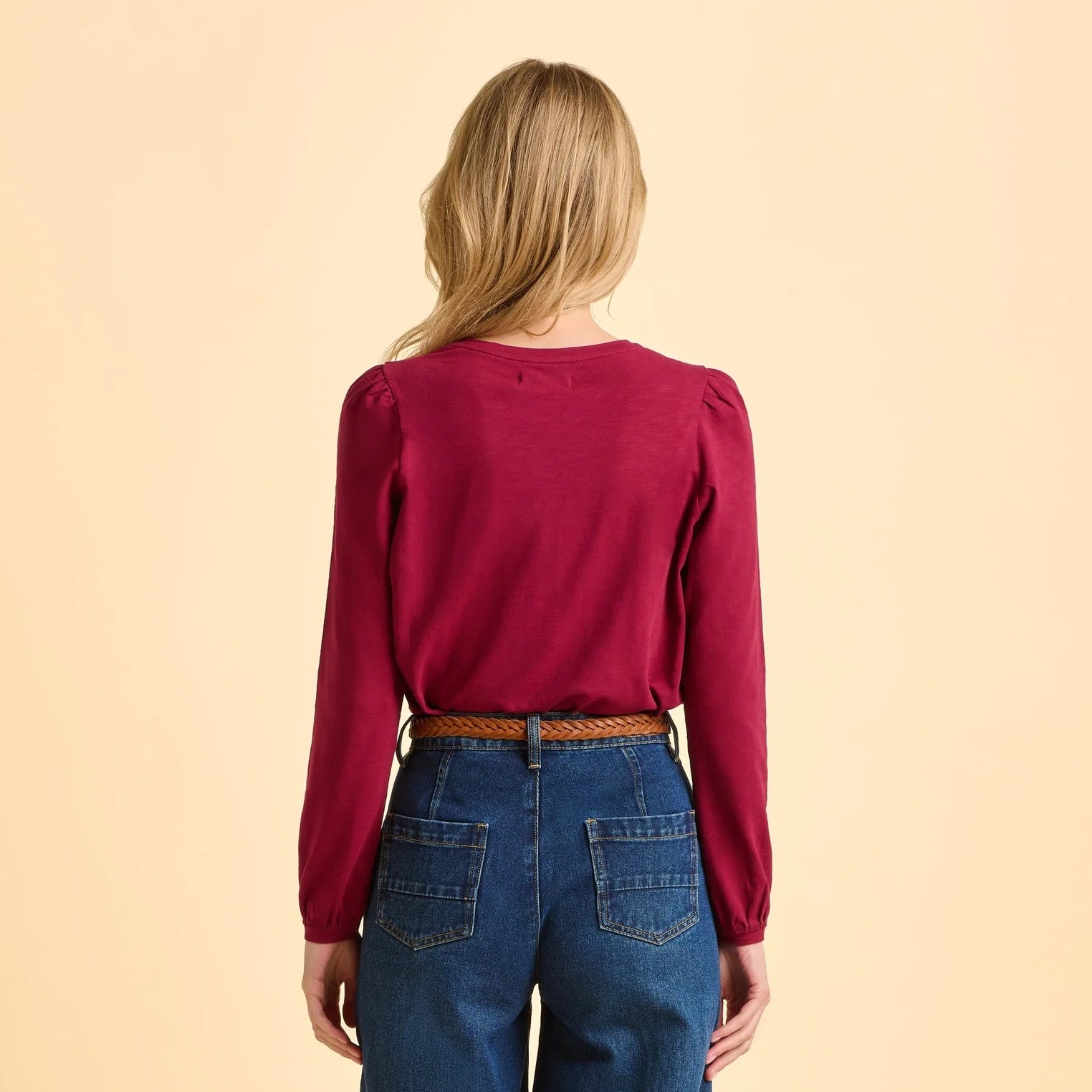 Back view of a woman wearing a burgundy red Brakeburn Janis long-sleeve t-shirt and blue jeans on a beige background