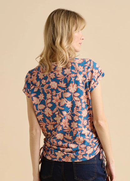 Back view of a woman wearing a Brakeburn short sleeve Midnight Garden short sleeve t-shirt in navy blue with a red and cream floral pattern and tie side detail, on a beige background