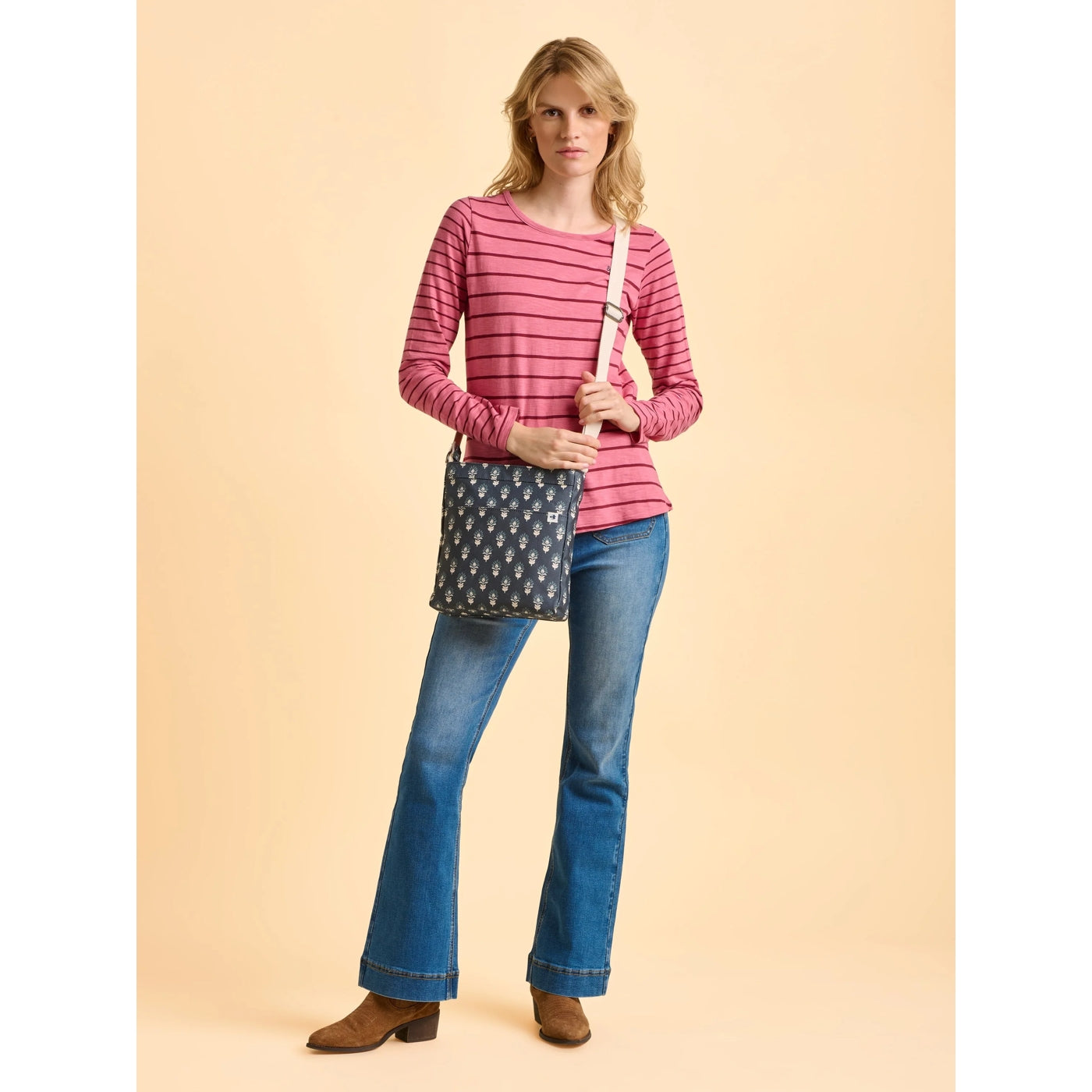 Woman wearing a pink striped shirt and blue jeans, holding a floral patterned Brakeburn Henna floral cross body bag on a beige background