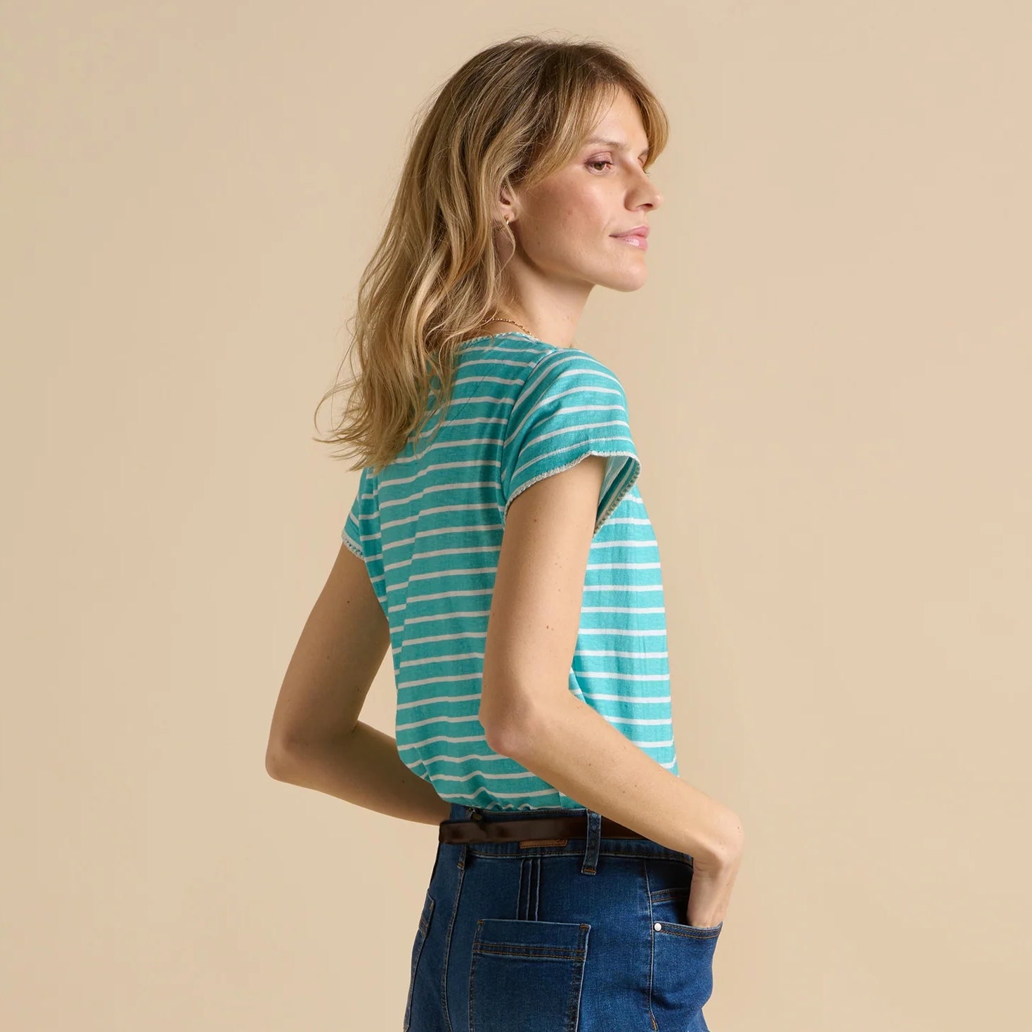 Side view of a woman wearing a short sleeve Brakeburn aqua and white stripe tee with jeans, on a beige background