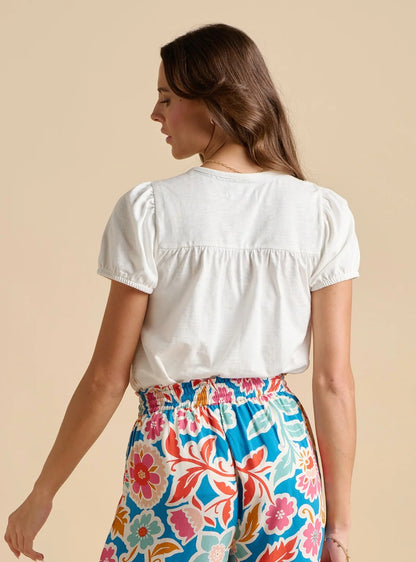 Back view of a woman wearing a Brakeburn short sleeve white Suri blouse with multicoloured floral trousers on a beige background