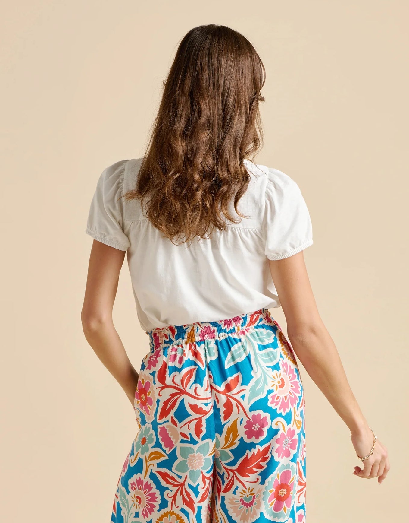 Back view of a woman wearing a white short sleeve Brakeburn Sui blouse with multicoloured trousers on a beige background
