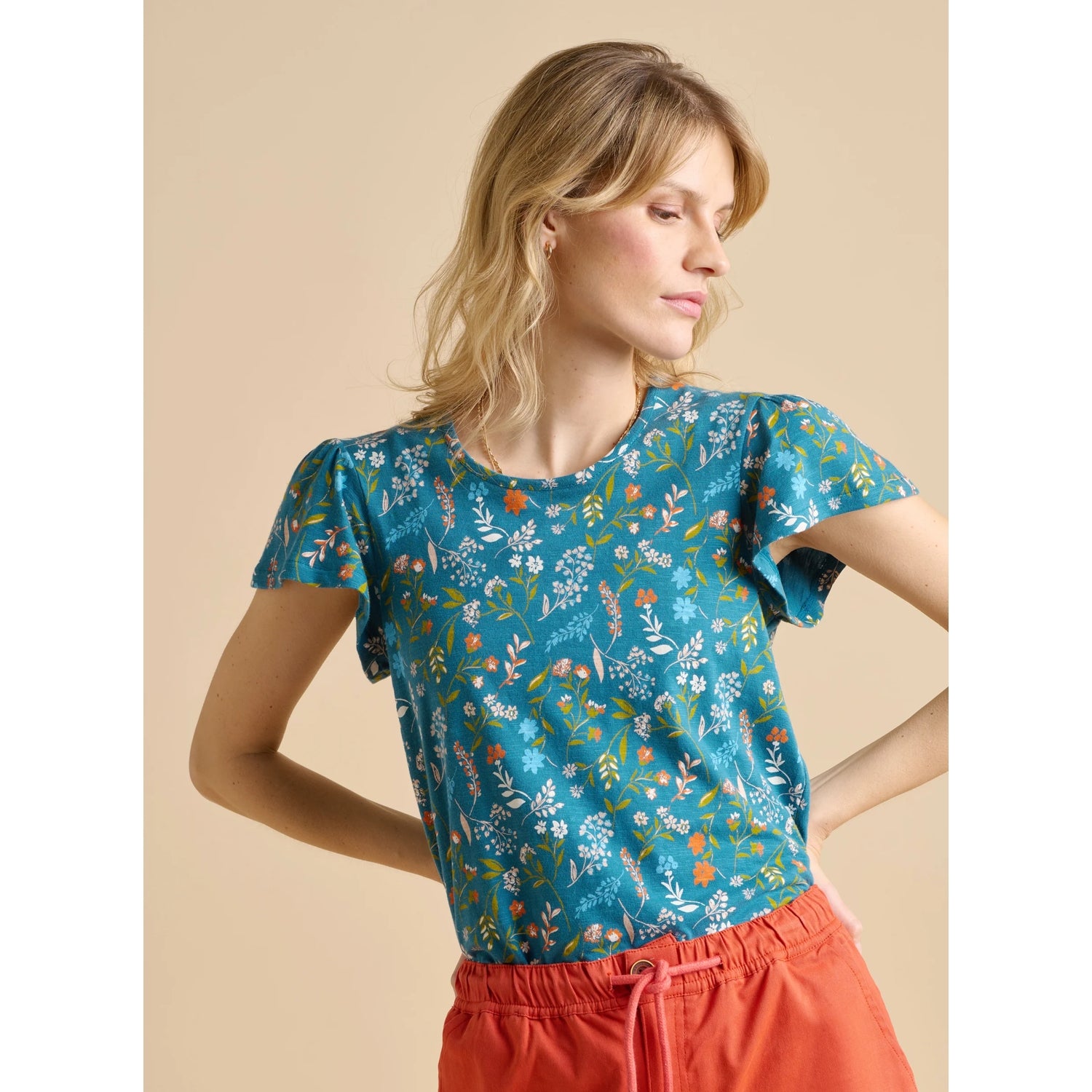 Woman wearing a Brakeburn teal meadow floral top and red pants on a beige background