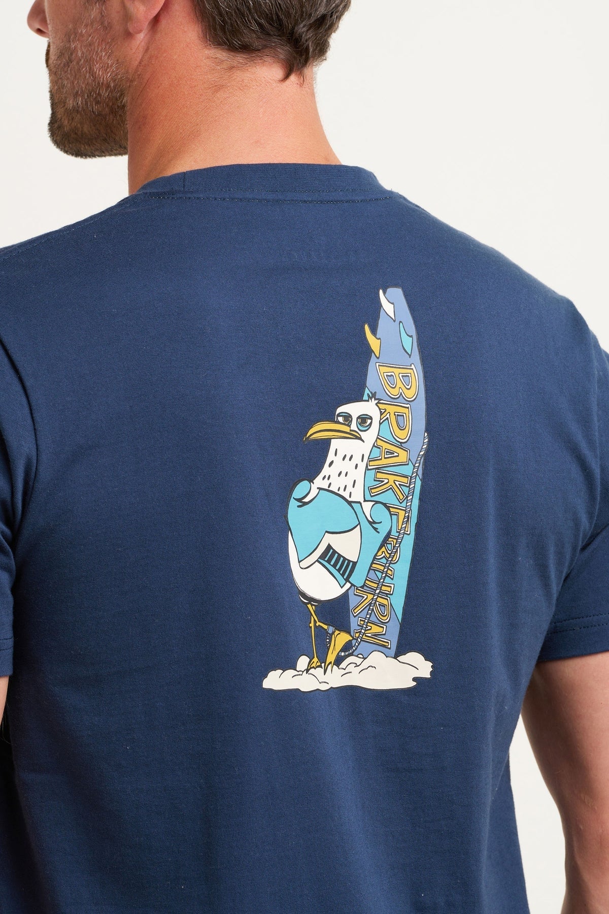 A men's tee from Brakeburn in navy featuring a print of a seagull with surfboard on the back and a smaller version on the front.