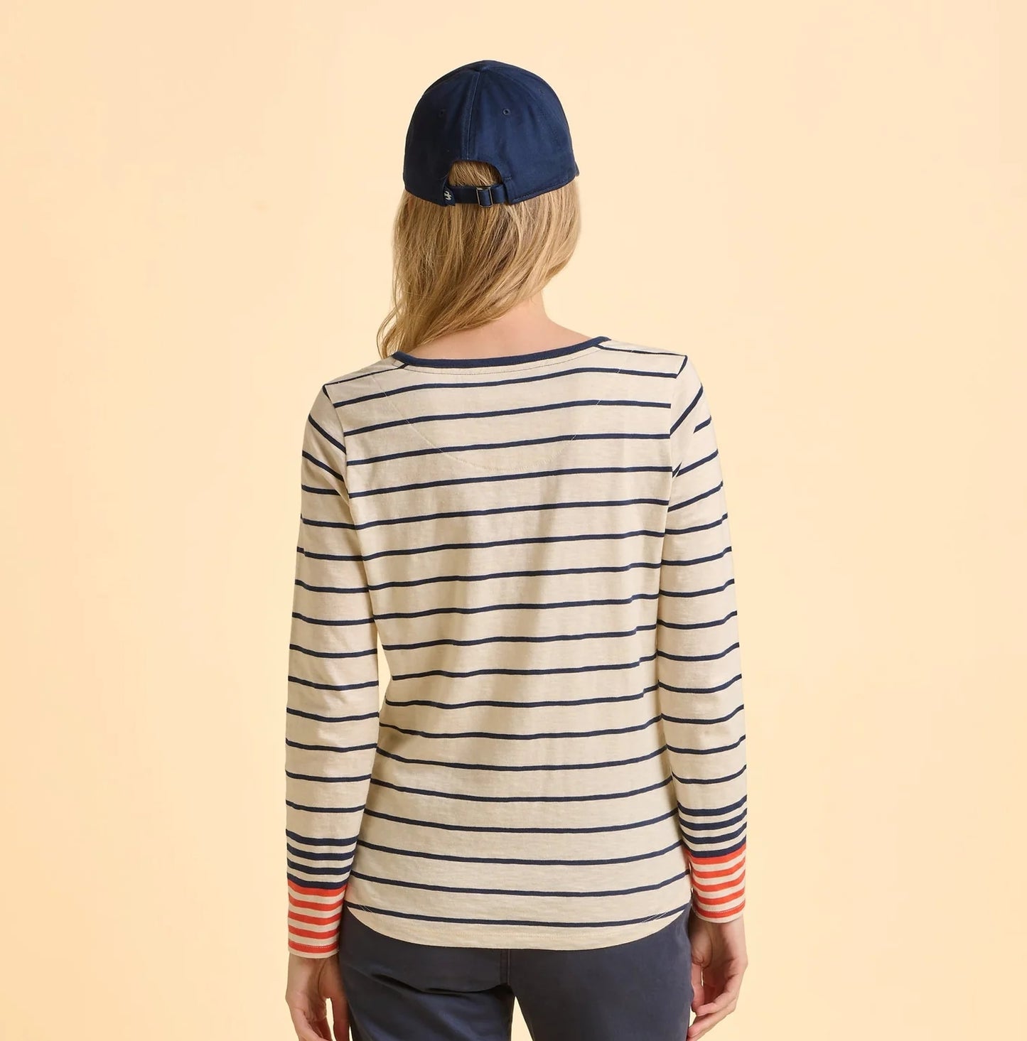 Back view of a woman wearing a striped Brakeburn long-sleeve Knoll t-shirt and navy cap on a beige background