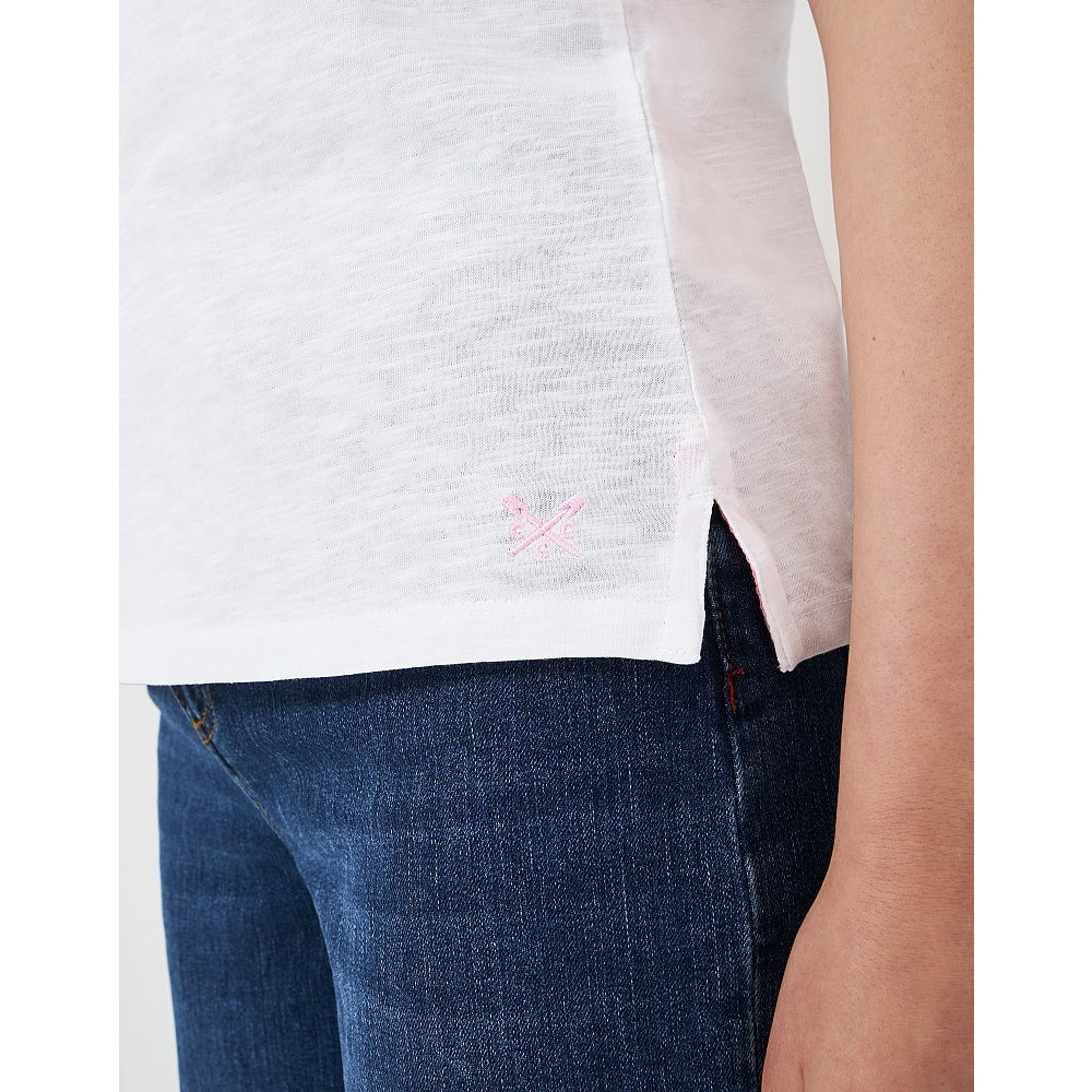 Close-up view of a logo and side seam slit of a womens Crew Clothing Perfect short sleeve plain t-shirt in white, with blue jeans and a red jumper