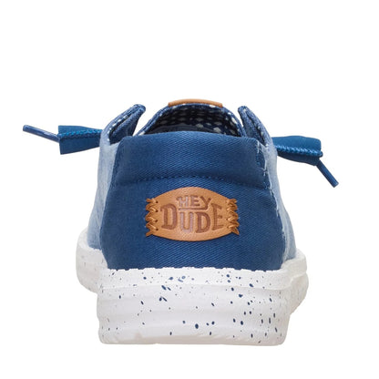 Back heel view of a women's Dude Wendy Chambray Grid shoe in Ensign Blue, featuring a speckled white sole and Hey Dude logo on the heel.