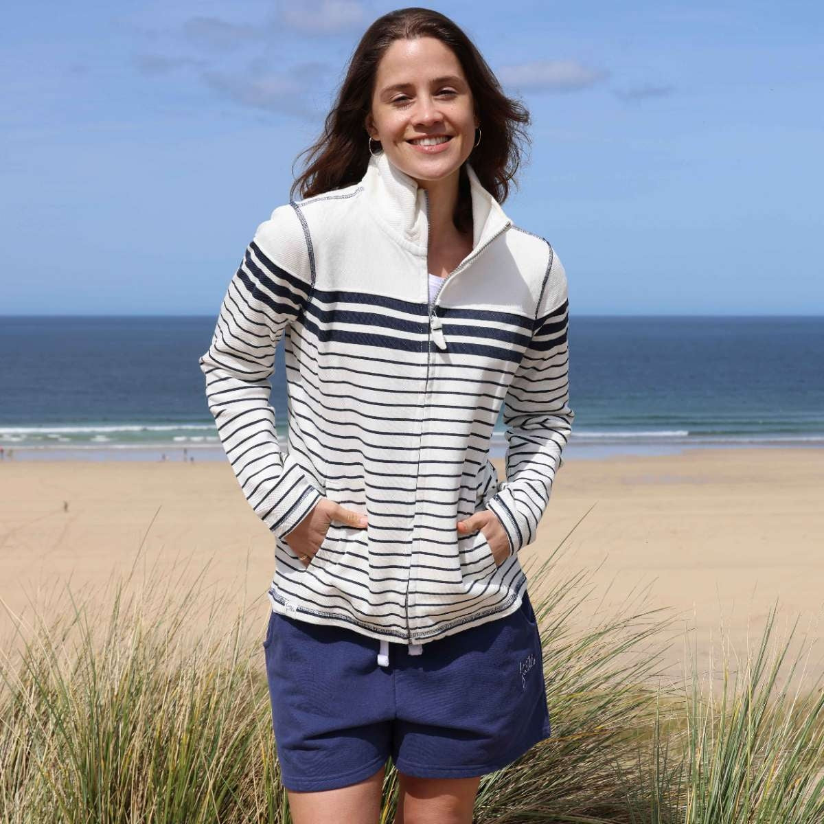 A woman wearing a Lazy Jacks LJ304S full zip stripe sweatshirt in off white with Twilight navy stripes and blue shorts.