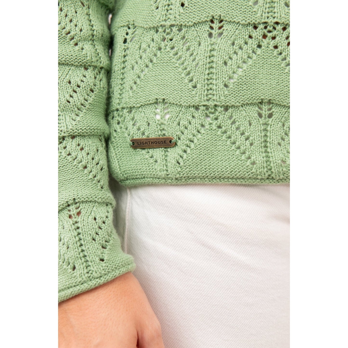 Close-up view of the geometric pointelle knit pattern and metal logo fob of a womens Lighthouse Eden knitted jumper in Soft Sage Green.