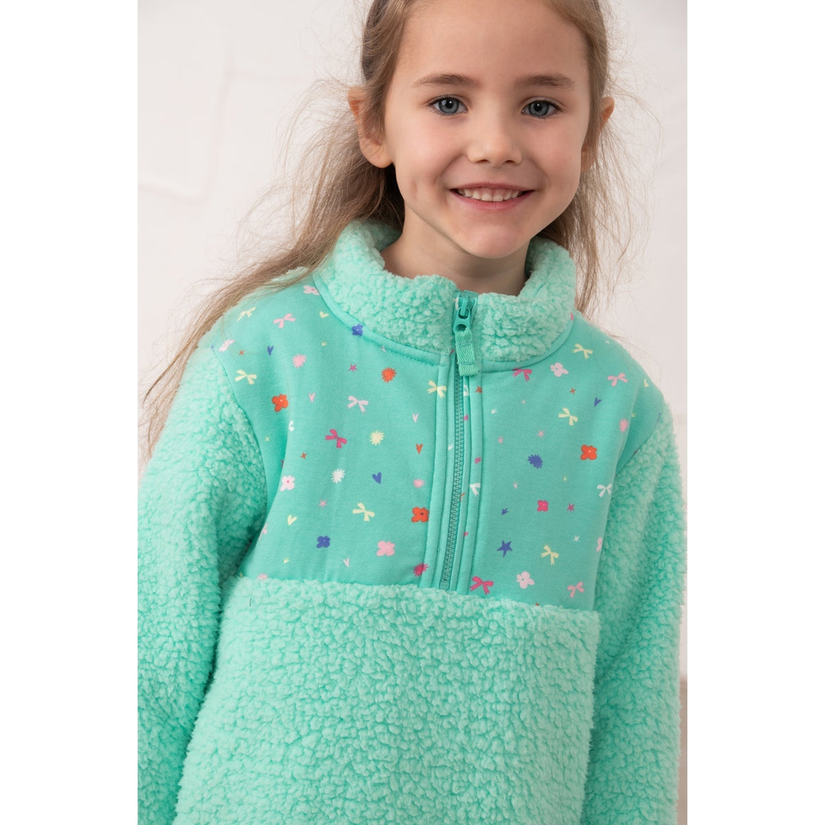 Close-up view of a girl wearing a Lighthouse Cassie fleece, in mint green with a tiffany bows, hearts, stars and floral print on the chest.