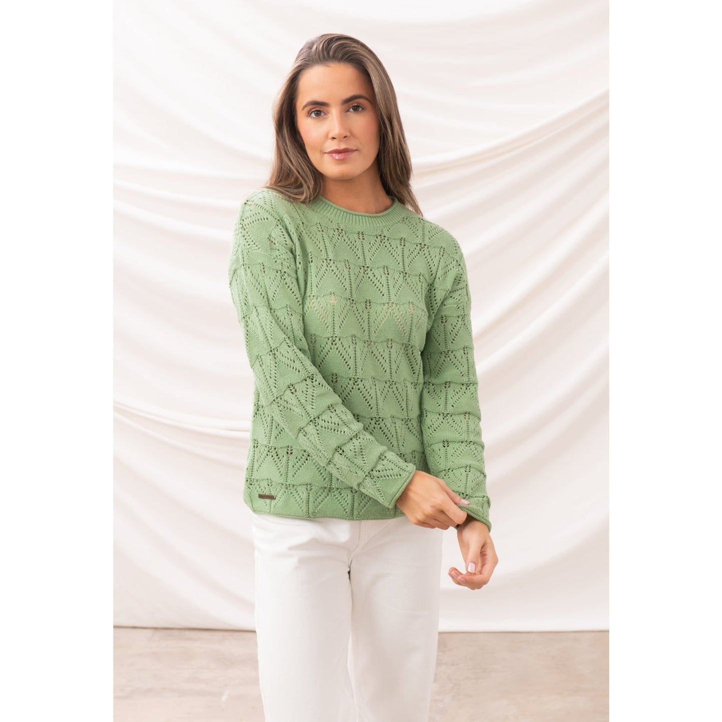 Front view of a woman wearing a Lighthouse Eden knitted jumper in Soft Sage Green with a geometric pointelle knit pattern and white trousers.