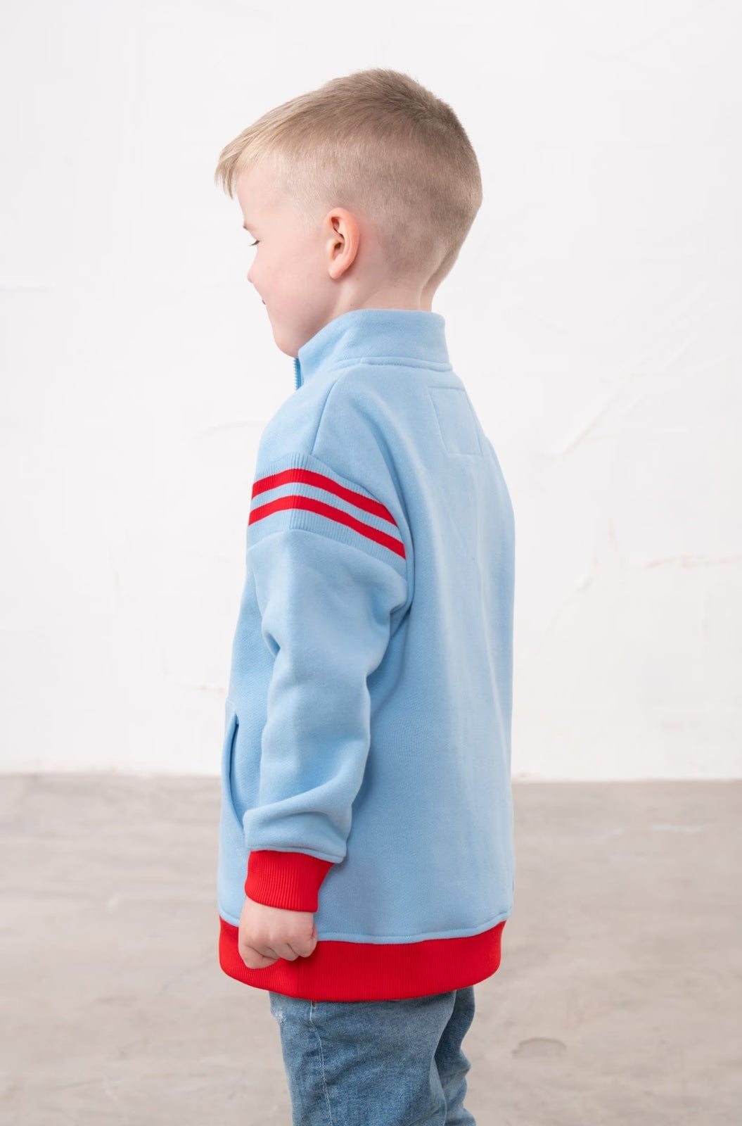 Back view of a boy wearing a Lighthouse Jonah 1/4 zip sweatshirt in sky blue with red trim and a red embroidered tractor applique on the chest.