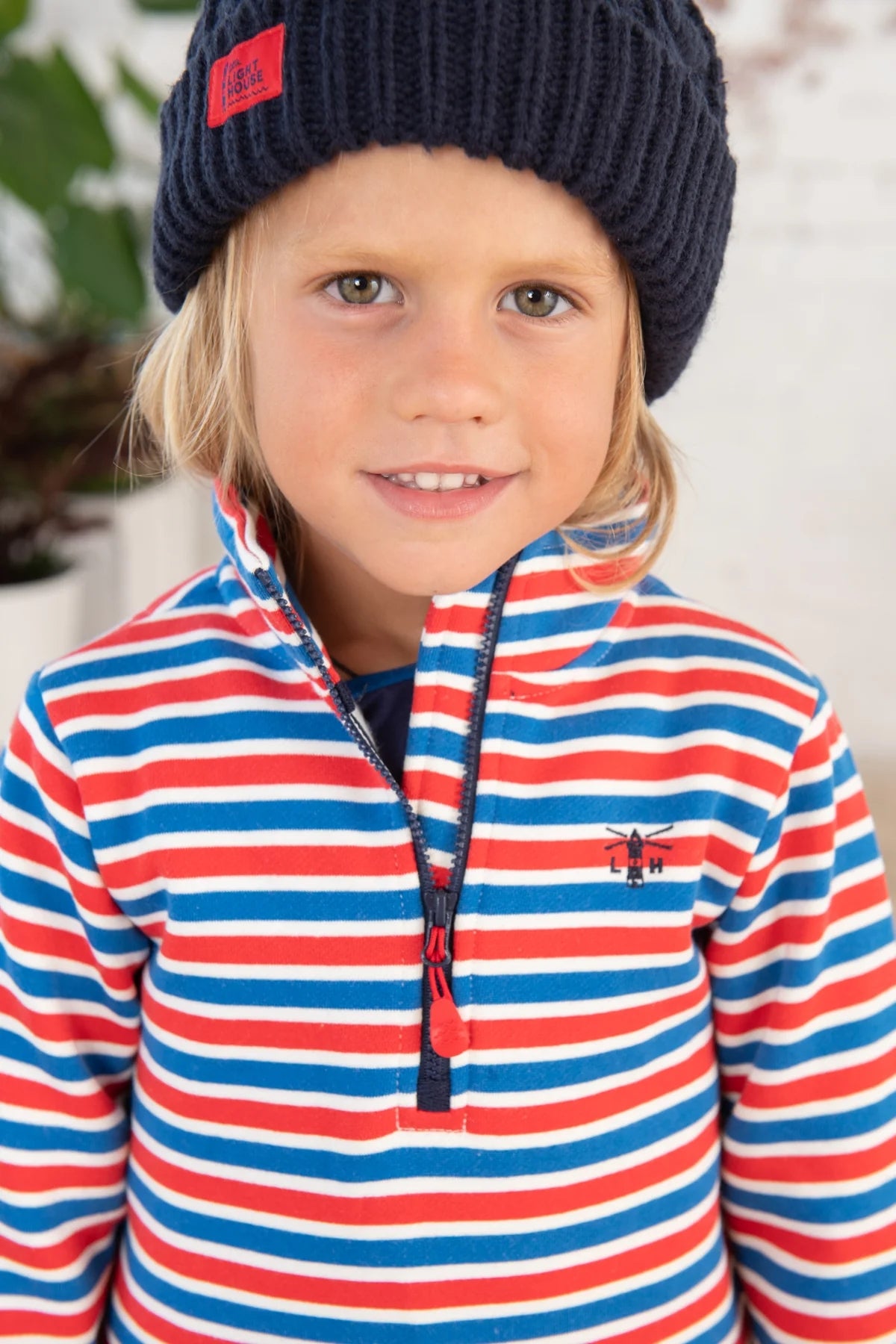 Closeup of a kid wearing a red, blue and white stripe Lighthouse Riley sweatshirt with a 1/4 zip neckline and navy beanie hat.