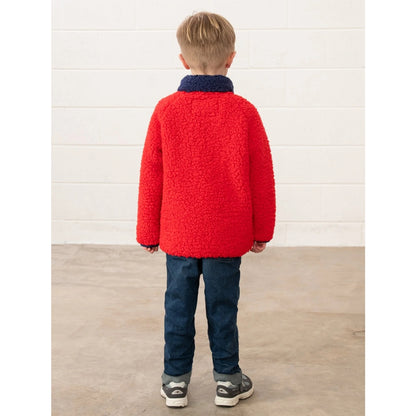 Back view of a child wearing a Lighthouse Caden red jumper and blue pants standing against a white wall.
