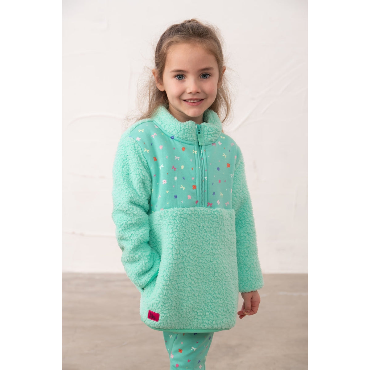 Front view of a girl wearing a Lighthouse Cassie fleece, in mint green with a tiffany bows, hearts, stars and floral print on the chest with side pockets.