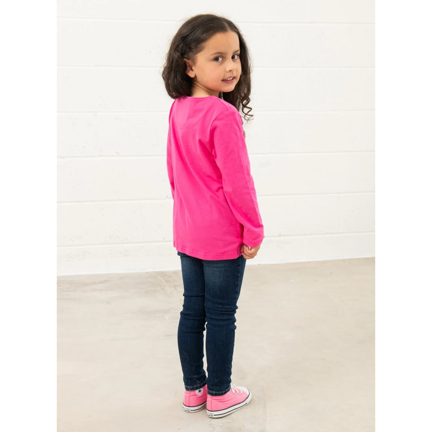 Back view of a young girl wearing a Lighthouse bright pink long-sleeve Causeway tee and dark blue jeans against a white background