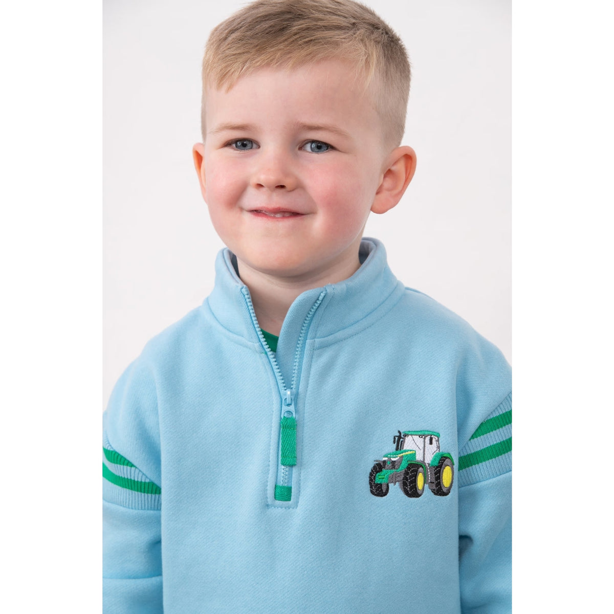 Close-up view of a child wearing a Lighthouse Jonah zip neck sweatshirt in sky blue with green trim and tractor applique on the chest.