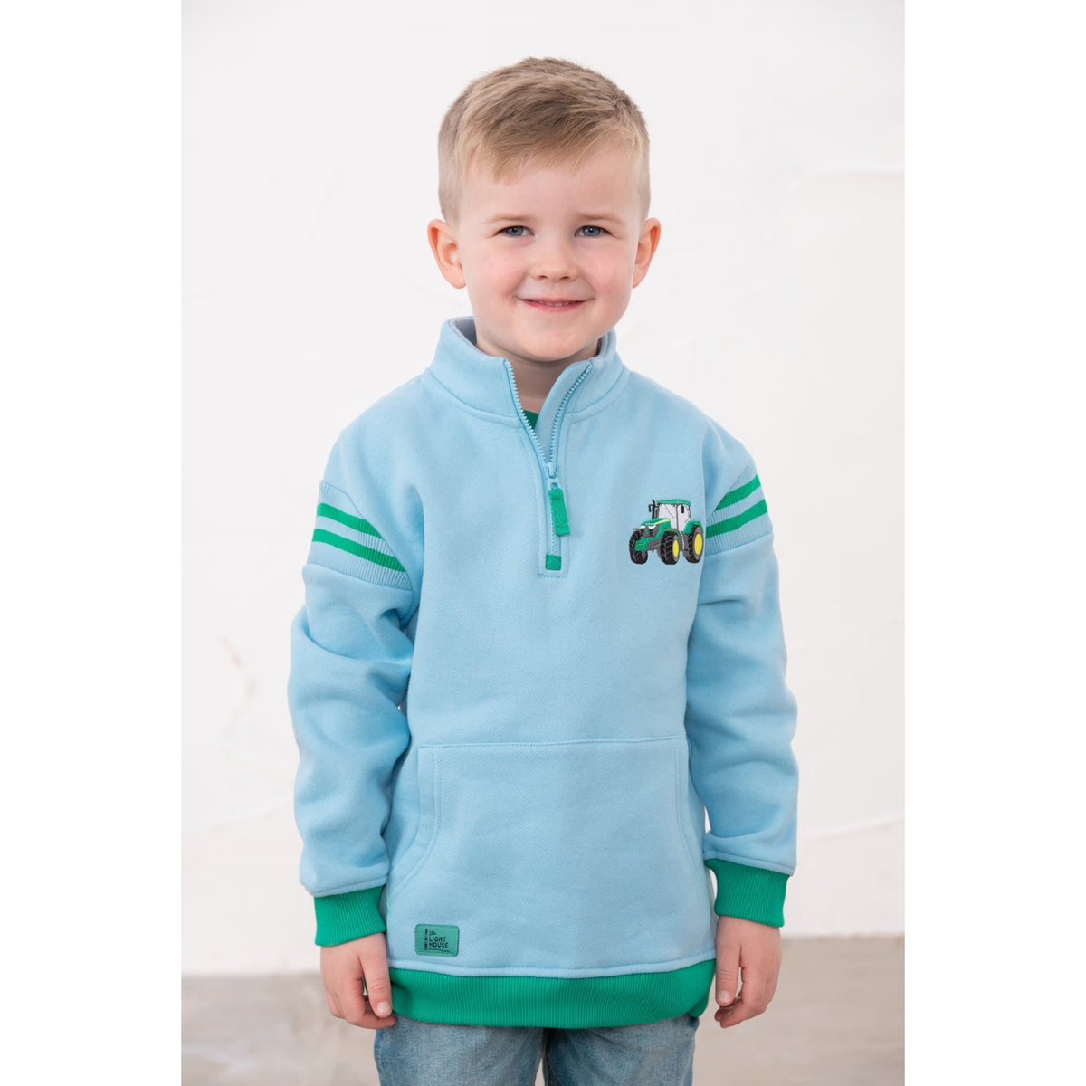 Front view of a child wearing a Lighthouse Jonah zip neck sweatshirt in sky blue with green trim and tractor applique on the chest.